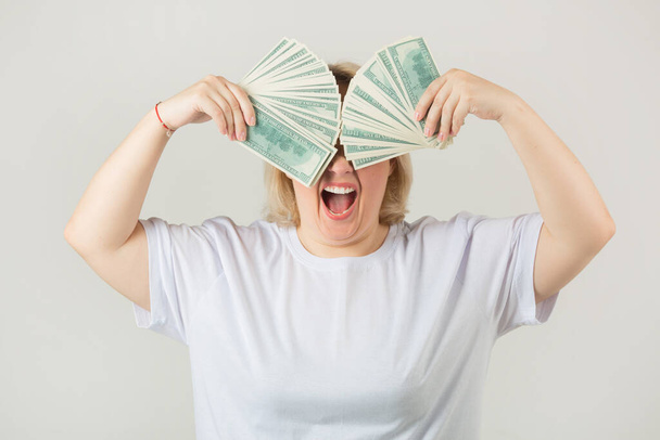beautiful young plump woman in a white t-shirt on a white background with dollars in her hands - Foto, imagen