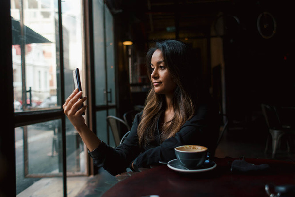 Młoda mieszana dziewczyna wyścig biorąc selfie ze smartfonem i siedzi przy stole z kawą  - Zdjęcie, obraz