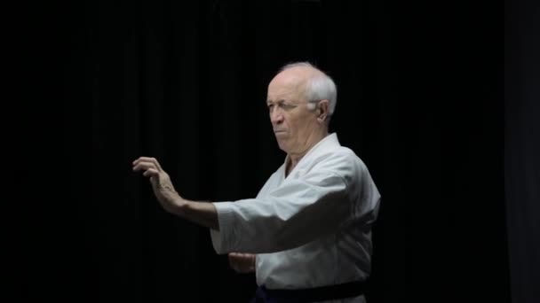 Sobre un fondo negro en karategi, un viejo atleta está entrenando golpes y bloqueos con sus manos - Metraje, vídeo