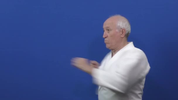 Sobre un fondo azul en karategi, un viejo atleta está entrenando golpes y bloqueos con sus manos - Metraje, vídeo