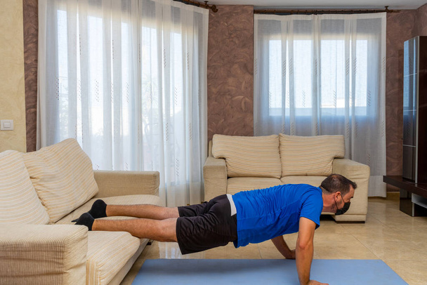 Homme barbu en forme basse faisant de l'exercice avec des vêtements de sport noirs et bleus et un masque pour prévenir le coronavirus dans le salon à la maison sur un tapis - Photo, image