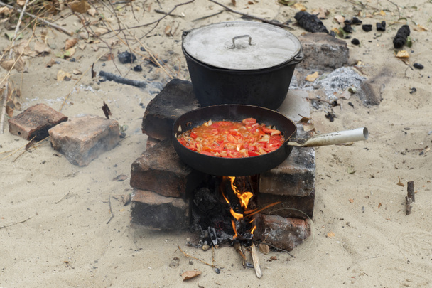 czarna żelazna patelnia z pieczonymi warzywami i duży garnek gotowania z stoiskiem zupy w ognisku i przygotowuje się do kolacji na świeżym powietrzu. Camping piknik - Zdjęcie, obraz
