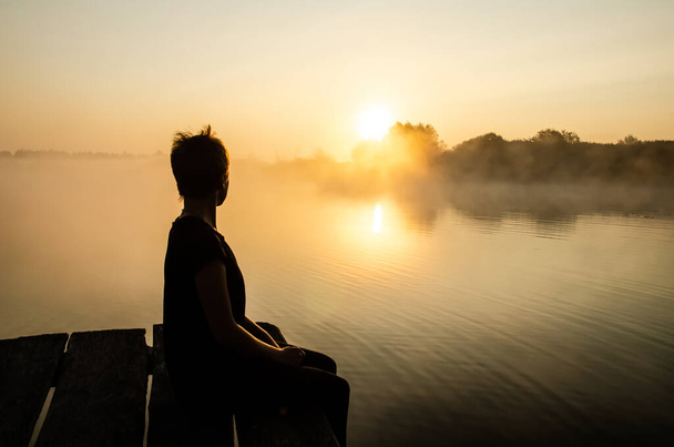 コピースペースと日没の空に対して桟橋に座っている少女のシルエット。少女は霧の中で湖の上に昇る太陽を見ている。水面の太陽の反射. - 写真・画像