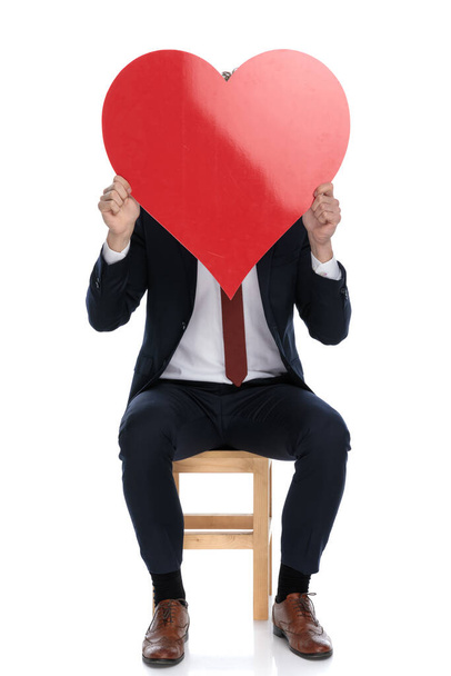 businessman hiding his face behind a big red heart and sitting on white background - Fotoğraf, Görsel