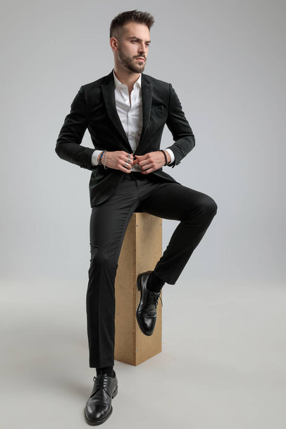 ελκυστικό μοντέλο μόδας ξεκουμπώνει το σακάκι του, κοιτάζοντας μακριά και κάθεται πάνω σε γκρι φόντο - Φωτογραφία, εικόνα