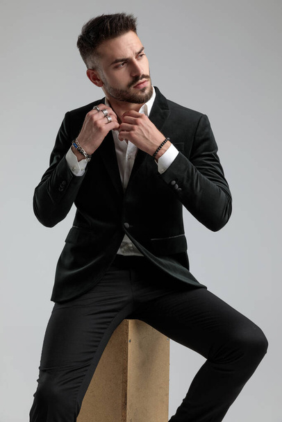 cool sexy businessman fixing his collar while looking aside and sitting against gray background - Foto, Imagem