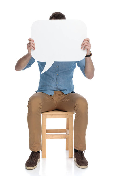 Joven hombre casual inteligente que cubre su cara con burbuja del habla mientras está sentado en una silla en el fondo del estudio blanco - Foto, Imagen