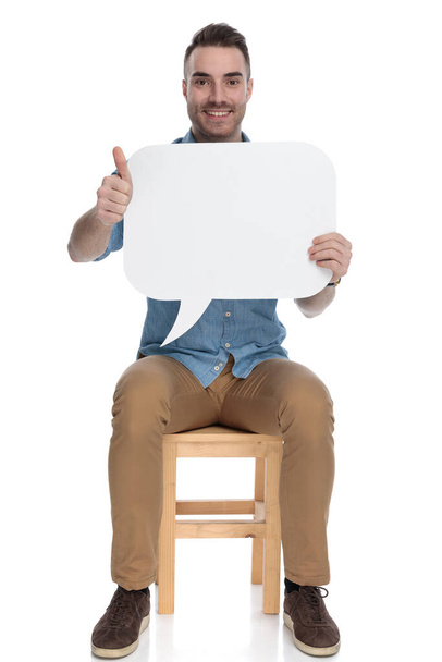 Happy smart casual man holding speech bubble and gesturing ok while sitting on a chair on white studio background - Photo, Image