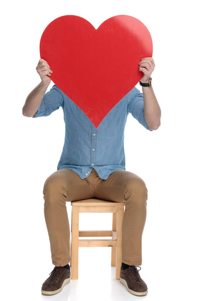 jong slim casual man bedekken zijn gezicht met hart vorm terwijl zitten op een stoel op witte studio achtergrond - Foto, afbeelding