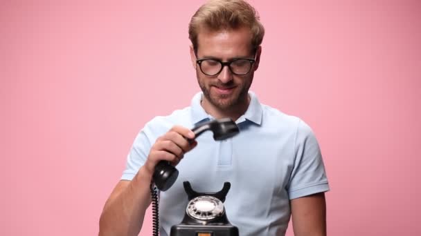 laughing young casual guy in blue polo shirt with eye glasses answering old rotary telephone, nodding, smiling and making thumbs up gesture on pink background - Footage, Video