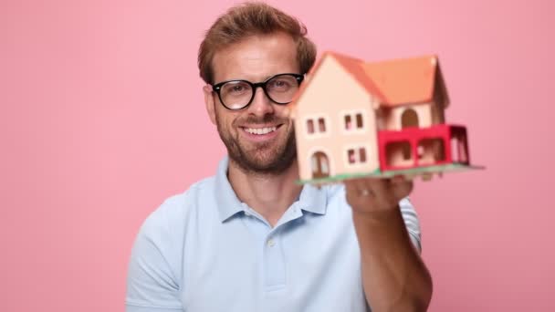 青いポロシャツを着た若いカジュアルな幸せな男が家の模型を見せ笑顔で親指を立てピンクの背景にうなずき - 映像、動画