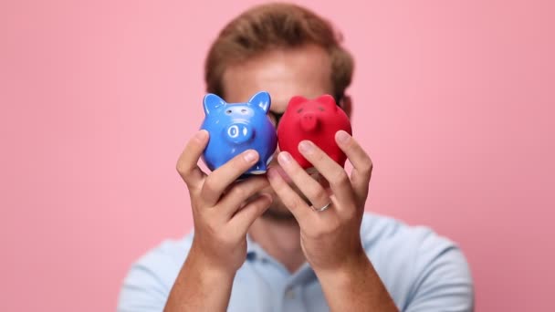 happy young casual man in blue polo shirt holding piggy banks in front of face, nodding and smiling on pink background - Footage, Video