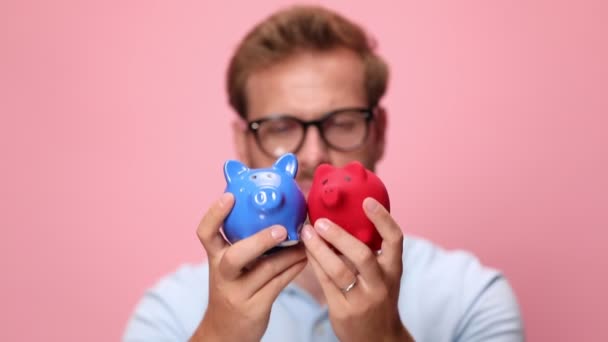 happy young casual man in blue polo holding piggy banks in front of face, analysing, nodding and smiling on pink background - Footage, Video