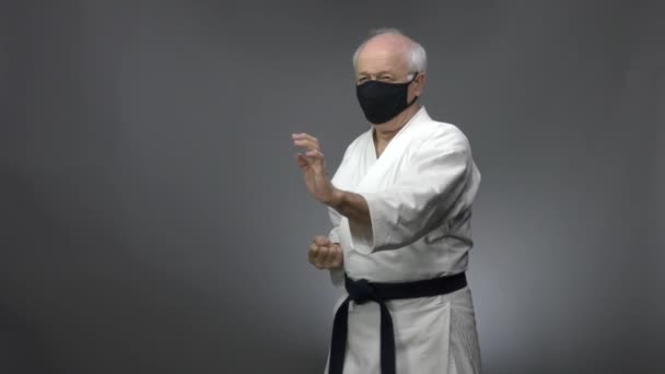 Em um fundo cinza, um velho atleta em uma máscara médica preta treina blocos e socos de braços - Filmagem, Vídeo
