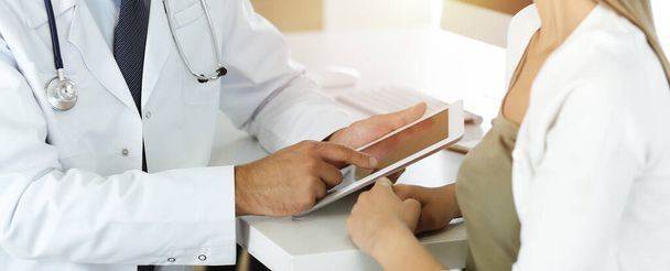Doctor and patient discussing medical exam results while sitting at the desk in sunny clinic, close-up. Male physician using tablet computer for filling up medication history record. - 写真・画像
