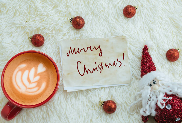top view red cup of cappuccino and Santa Claus toy on a background of fluffy white blanket   with inscription merry Christmas on a vintage paper - Foto, Bild