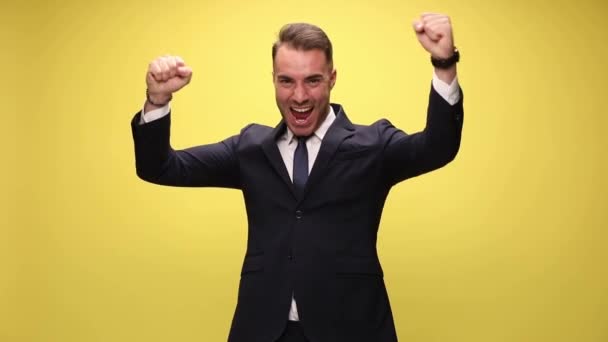 fiatal jóképű üzletember felemeli mindkét öklét, és ünnepli sikerek sárga háttér - Felvétel, videó