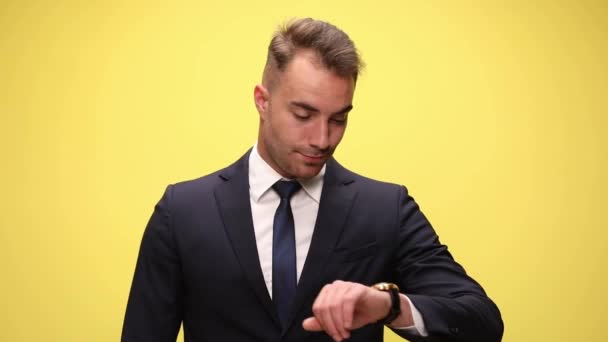 joven hombre de negocios guapo comprobando el tiempo en su reloj, siendo sorprendido y apuntando a su cabeza en el fondo amarillo - Metraje, vídeo