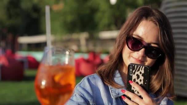 A woman drinks an orange cocktail and takes photos of it on her smartphone - Filmagem, Vídeo