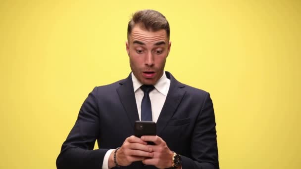 atractivo hombre de negocios mensajes de texto en su teléfono, siendo sorprendido, golpeando su cara y agitando la cabeza sobre el fondo amarillo - Metraje, vídeo