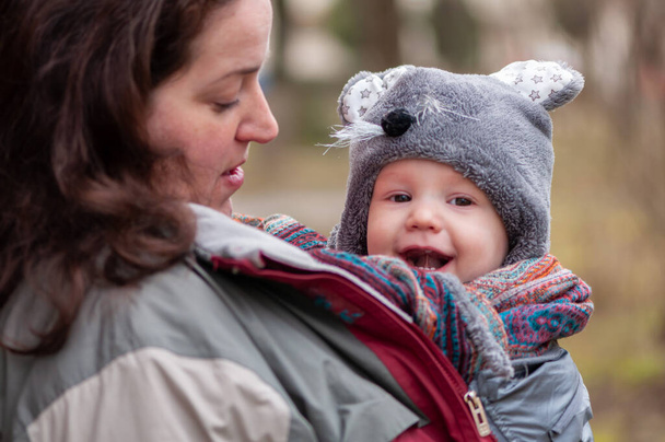 Portret van een moeder en haar baby. Moeder houdt haar baby worm met haar jas. Baby in doek omwikkeld. Grappige hoed.. - Foto, afbeelding