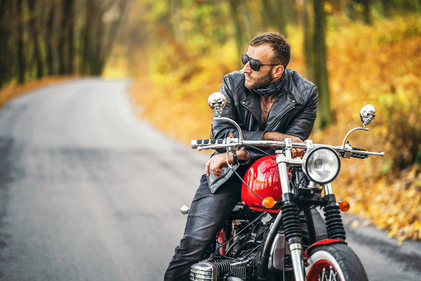 青白い背景を持つ森の道路上のオートバイに座ってサングラスや革のジャケットでひげを剃った残忍な男. - 写真・画像