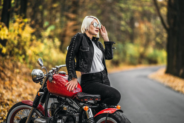 Pretty blonde biker girl in sunglasses with red motorcycle on the road in the forest with colorful blurred background. - Фото, изображение
