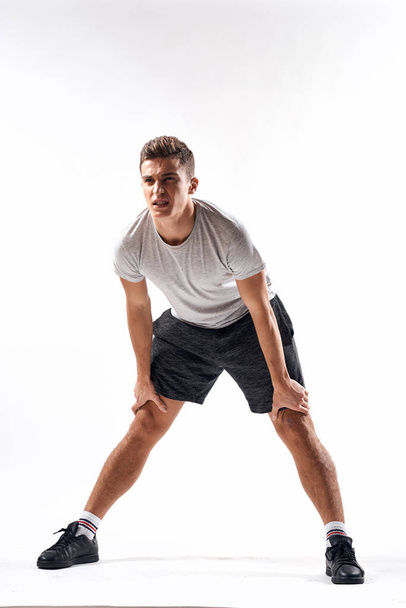 A sports man in shorts and a full-length T-shirt does exercises on a light background - Foto, imagen