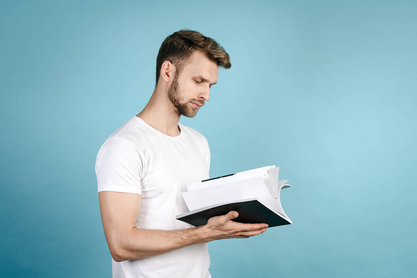 Вид сбоку на уверенного, молодого и умного студента, читающего научный журнал, стоящего изолированно на фоне синего копировального пространства в белой футболке. Концепция образования - Фото, изображение
