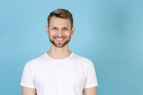 Portrait of happy young man wearing in white t-shirt, standing isolated on blue copy space background, looking at camera and smiling wide - Φωτογραφία, εικόνα