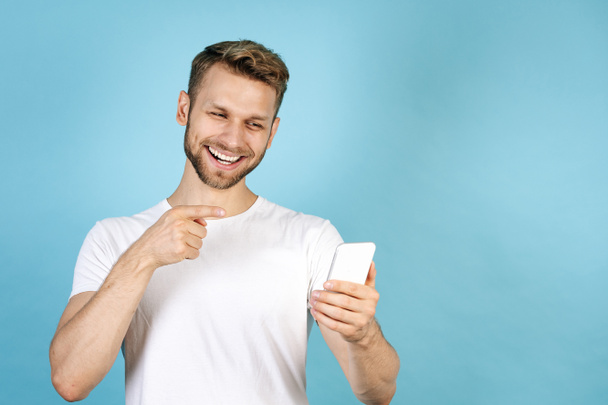 Ευτυχισμένος και χαμογελαστός νεαρός άνδρας με το δάχτυλο στην κάμερα στο σύγχρονο smartphone, κάνοντας online βιντεοκλήση, στέκεται απομονωμένος σε μπλε φόντο αντίγραφο χώρου σε casual λευκό t-shirt - Φωτογραφία, εικόνα