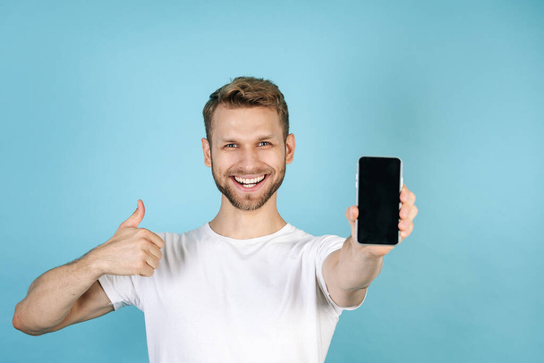 Koncept reklamy. Šťastný a usmívající se mladý muž v ležérní bílé tričko ukazující palce nahoru, držící moderní smartphone s prázdným displejem na obrazovce, stojící izolovaně na modrém pozadí kopírování - Fotografie, Obrázek