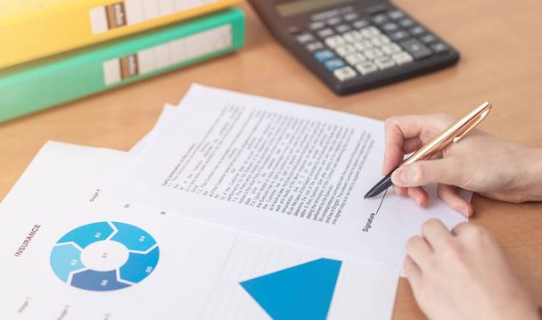 Businesswoman is signing a contract at office table with folders, calculator and financial documents with charts and contract, female hands - Photo, image