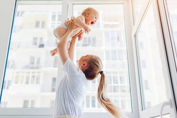 Mother playing with newborn baby son at home. Woman tossing kid up on balcony. Happy infant wearing diaper. Isolation during quarantine - Zdjęcie, obraz