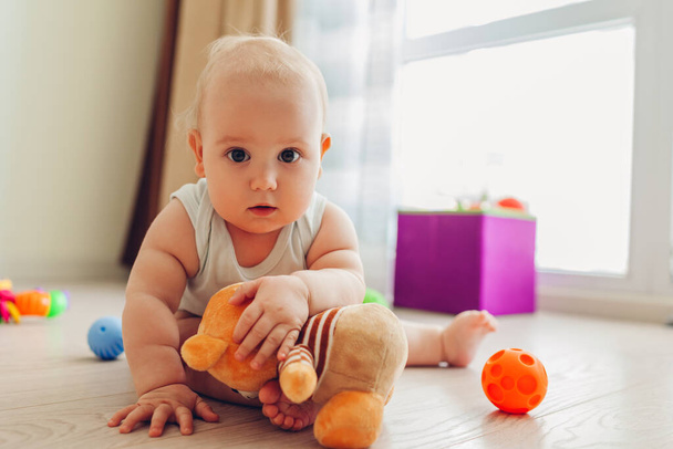 Little newborn baby playing with toys sitting on floor at home. Kid looking at camera holding teddy bear - Photo, Image