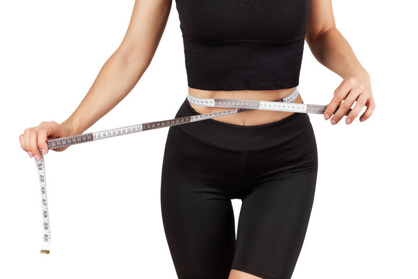 a young beautiful woman in a black tracksuit measures her waist with a centimeter tape, isolate on a white background - Photo, image