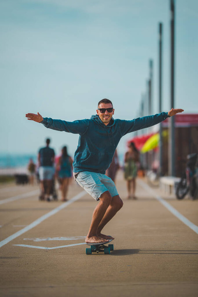 Glücklich unbeschwerter Skater in Kapuzenpulli und Sonnenbrille fährt auf einem Longboard. Konzept für Sommerspaß. - Foto, Bild