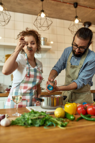 Italian man adding pepper, spice to the soup while woman tasting it. Couple preparing a meal together in the kitchen. Cooking at home, Italian cuisine - 写真・画像