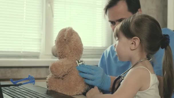 paciente criança senta-se no colo do pediatra. Cuidar profissional pediatra do sexo masculino brincando com uma criança pequena no escritório. menina brinca com um ursinho de pelúcia brinquedo ouve ele com um estetoscópio. - Filmagem, Vídeo