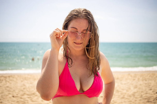 joven mujer caucásica juguetona en un bikini rosa y gafas corazones coquetamente mirando a la cámara. - Foto, Imagen