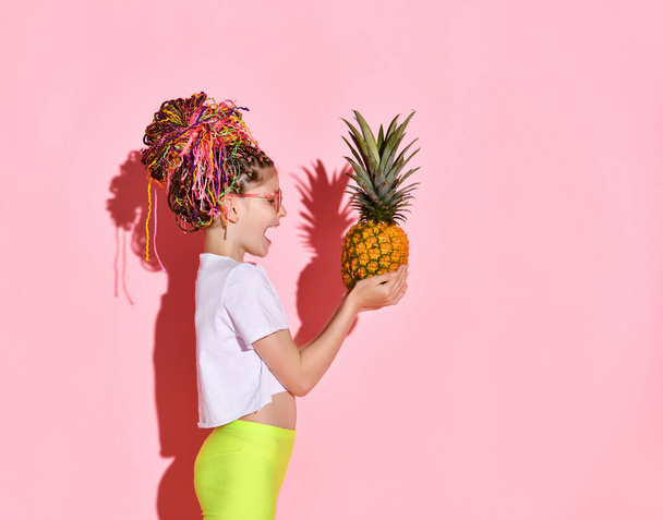 Side shot of an excited girl with colorful braids in stylish red glasses looking at a big pineapple in her hands. Waist shot isolated on pink, copy space. - Foto, Imagen