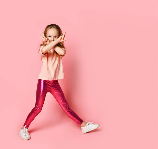 Cute little girl standing with legs wide apart and hiding her face behind two crossed hands. Full length shot isolated on pink, copy space - Foto, Imagem