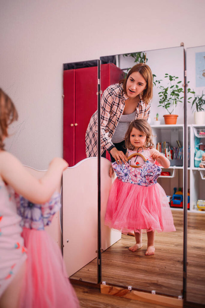 Holčička a maminka si vybírají oblečení. Dítě si zkouší šaty doma a dívá se do zrcadla - Fotografie, Obrázek