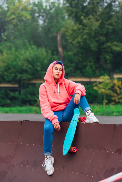 Young girl with short multicolored hair and nose piercing dressed in pink hoodie sits with skateboard in skatepark during morning fog. Portrait of a tomboy with plastic skateboard - Photo, Image