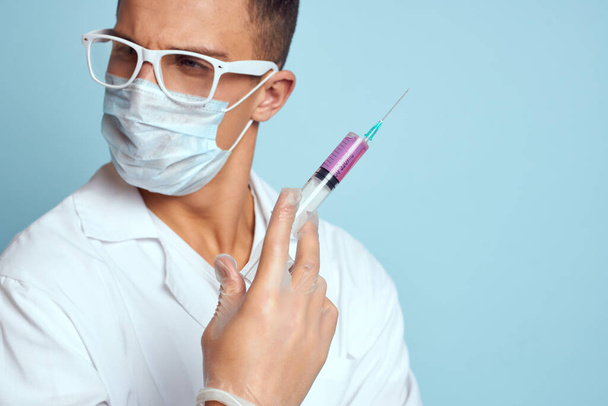 Ο γιατρός κρατά μια σύριγγα στο χέρι του με ένα κόκκινο υγρό σε ένα μπλε φόντο ιατρικό φόρεμα και μια προστατευτική μάσκα - Φωτογραφία, εικόνα