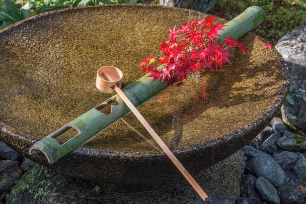 Πηγή νερού Tsukubai στο ιαπωνικό κήπο στο Zuiganzan Enkouji Temple, Κιότο, Ιαπωνία το φθινόπωρο. Με κόκκινα φύλλα σφενδάμου γύρω από το νιπτήρα. - Φωτογραφία, εικόνα