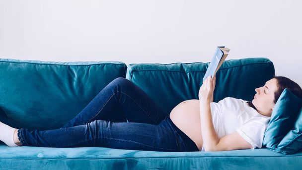 Домашній затишний портрет вагітної жінки, яка відпочиває вдома і читає книгу. Вагітна матір середнього віку портрет лежить на дивані вдома. Концепція здорової вагітності, брюнетка майбутня жінка. Копіювати простір
. - Фото, зображення
