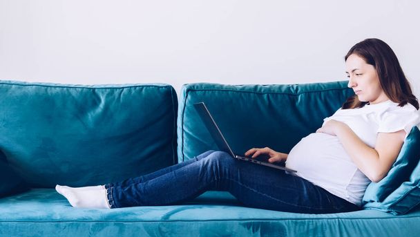 Young beautiful pregnant woman working on laptop lying on sofa. Pregnant business woman searching information and online shopping on laptop prepare for childbirth and parenting at home. - Foto, afbeelding