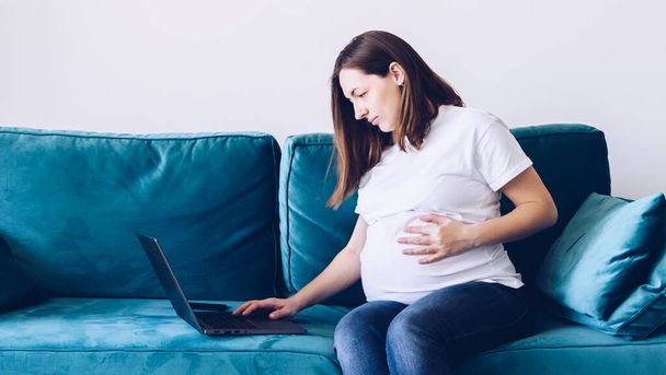 Молодая красивая беременная женщина, работающая на ноутбуке, лежит на диване. Беременная деловая женщина ищет информацию и онлайн-покупки на ноутбуке готовятся к родам и воспитанию дома. - Фото, изображение