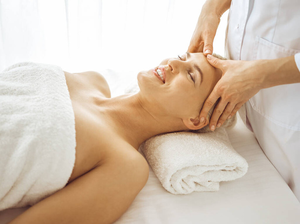 Beautiful blonde woman enjoying facial massage with closed eyes. Relaxing treatment in medicine and spa center concepts - Fotografie, Obrázek